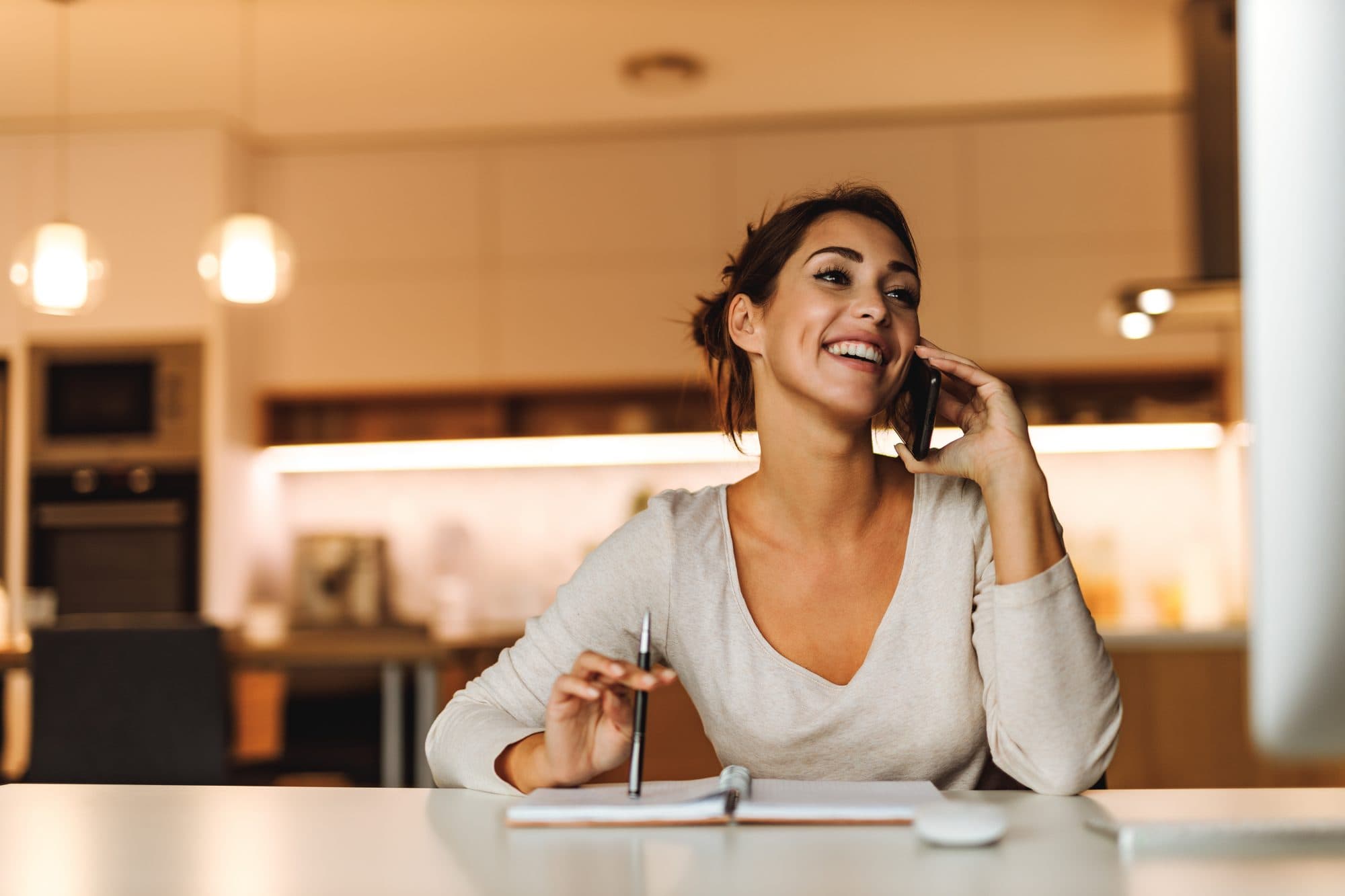 Smiling woman on a phone call at her kitchen table with a notebook and pen