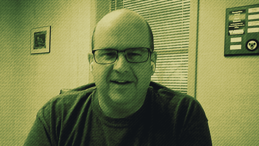 Bald man with glasses smiling in an office setting, green-tinted photo